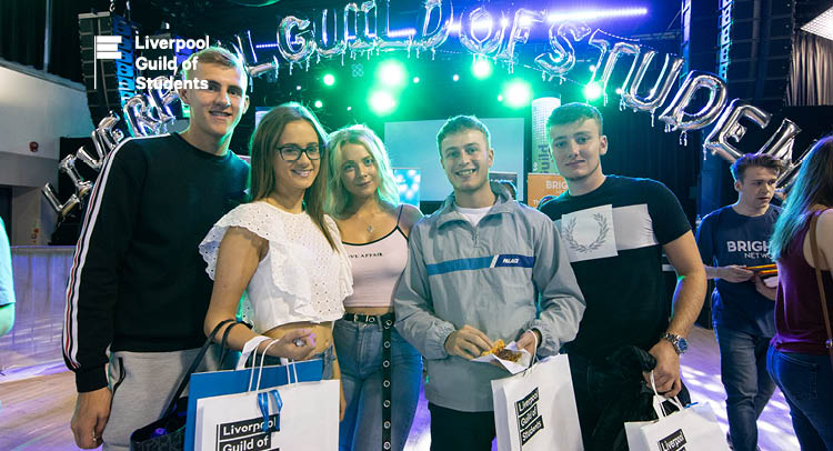 Group of 5 students holding Guild goody bags, standing in front of a balloon arch that reads Liverpool Guild of Students